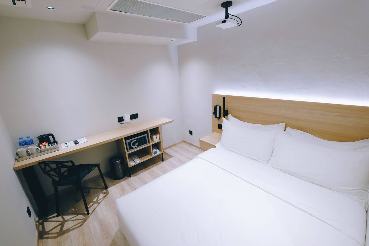 A hotel room with a bed, a desk, a chair, a microwave, a trash bin, and a nightstand with a lamp.