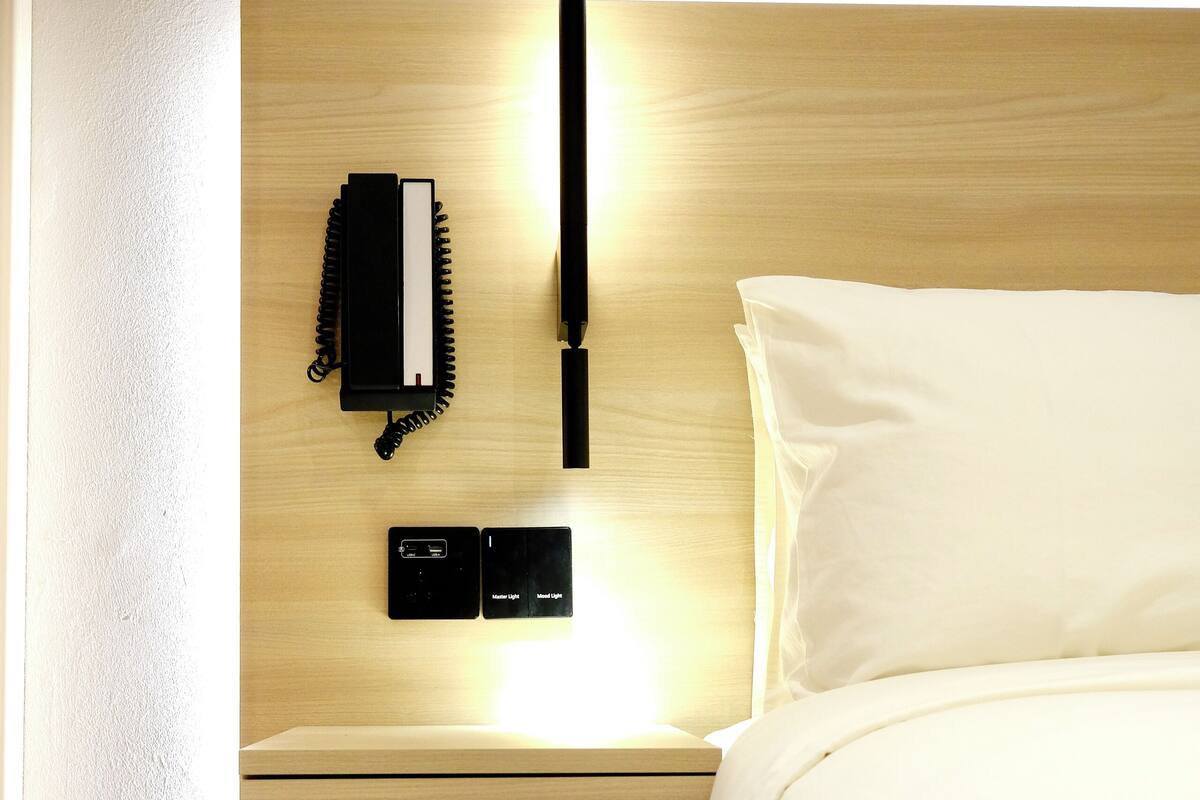 A neatly made bed with a wooden headboard, a bedside table with a phone and a small control panel, and a wall-mounted light.
