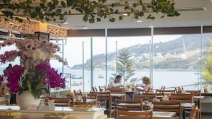 Ein Restaurant mit Holztischen und -stühlen, einer großen Blumenvase und Blick auf das Meer und die Hügel im Hintergrund.