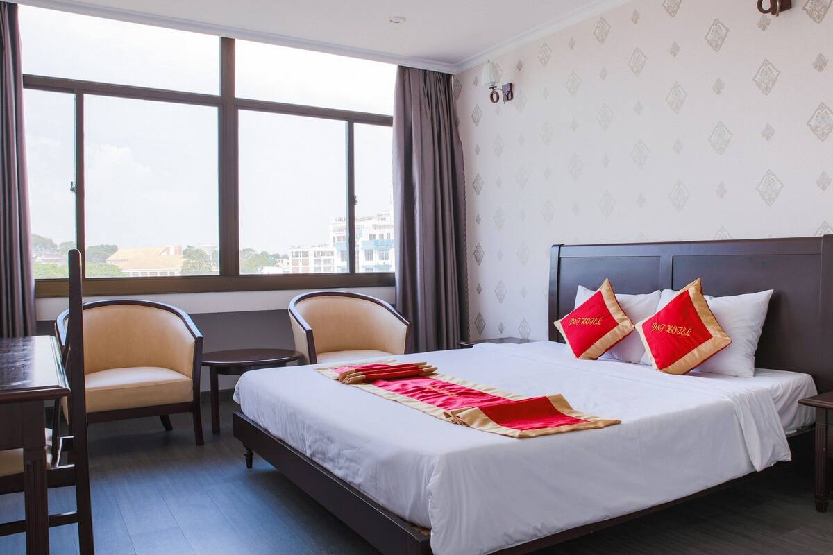 Une chambre d’hôtel avec un grand lit, deux chaises, une petite table et une fenêtre avec des rideaux.