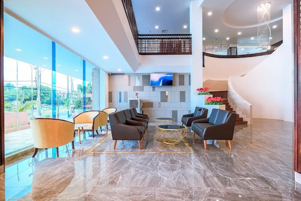A spacious lobby with a modern design, featuring a seating area with sofas and armchairs, a large flat-screen TV mounted on the wall, and a staircase leading to an upper level.