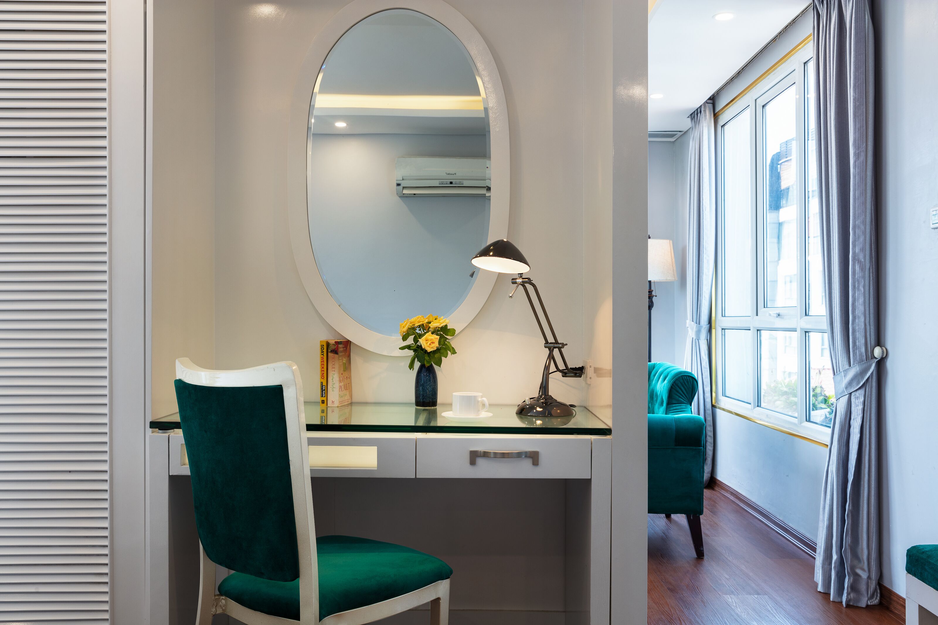 Una habitación moderna con escritorio, silla y un espejo grande. Hay una ventana con cortinas, una lámpara y un florero con flores sobre el escritorio.