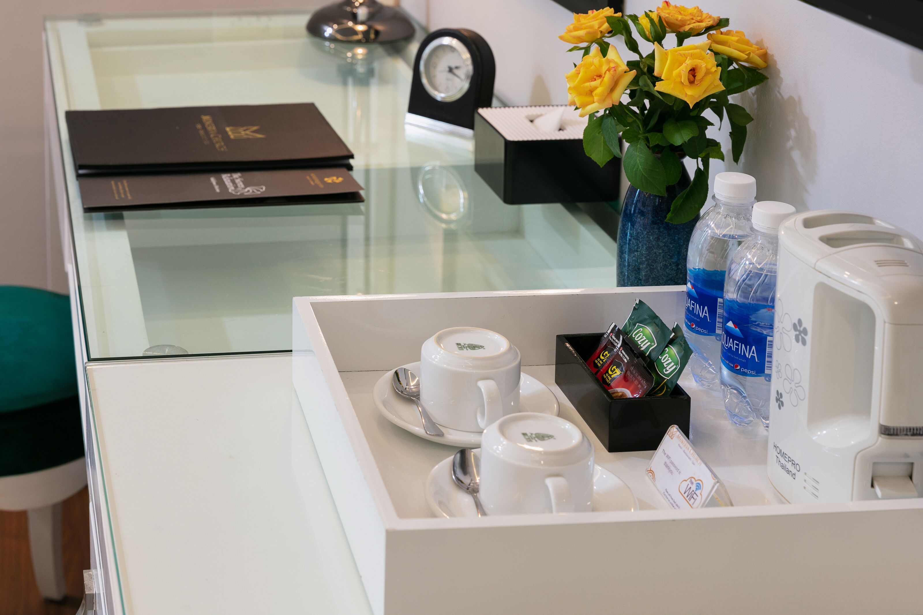 A hotel room's reception area with a glass partition, a clock, a vase with yellow flowers, and a tray with cups, a box of tissues, and bottled water.