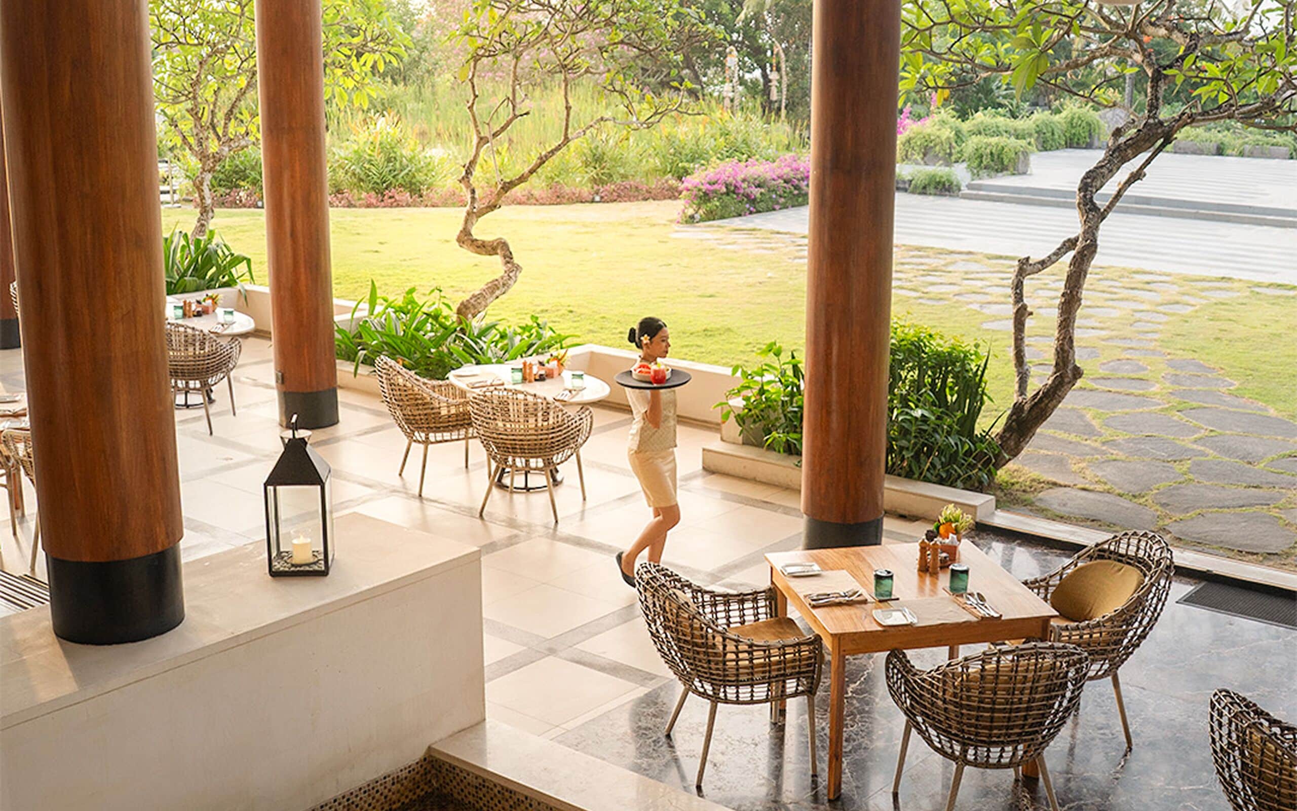 A restaurant with outdoor seating, wicker chairs, a waiter serving a dish, and a garden view.
