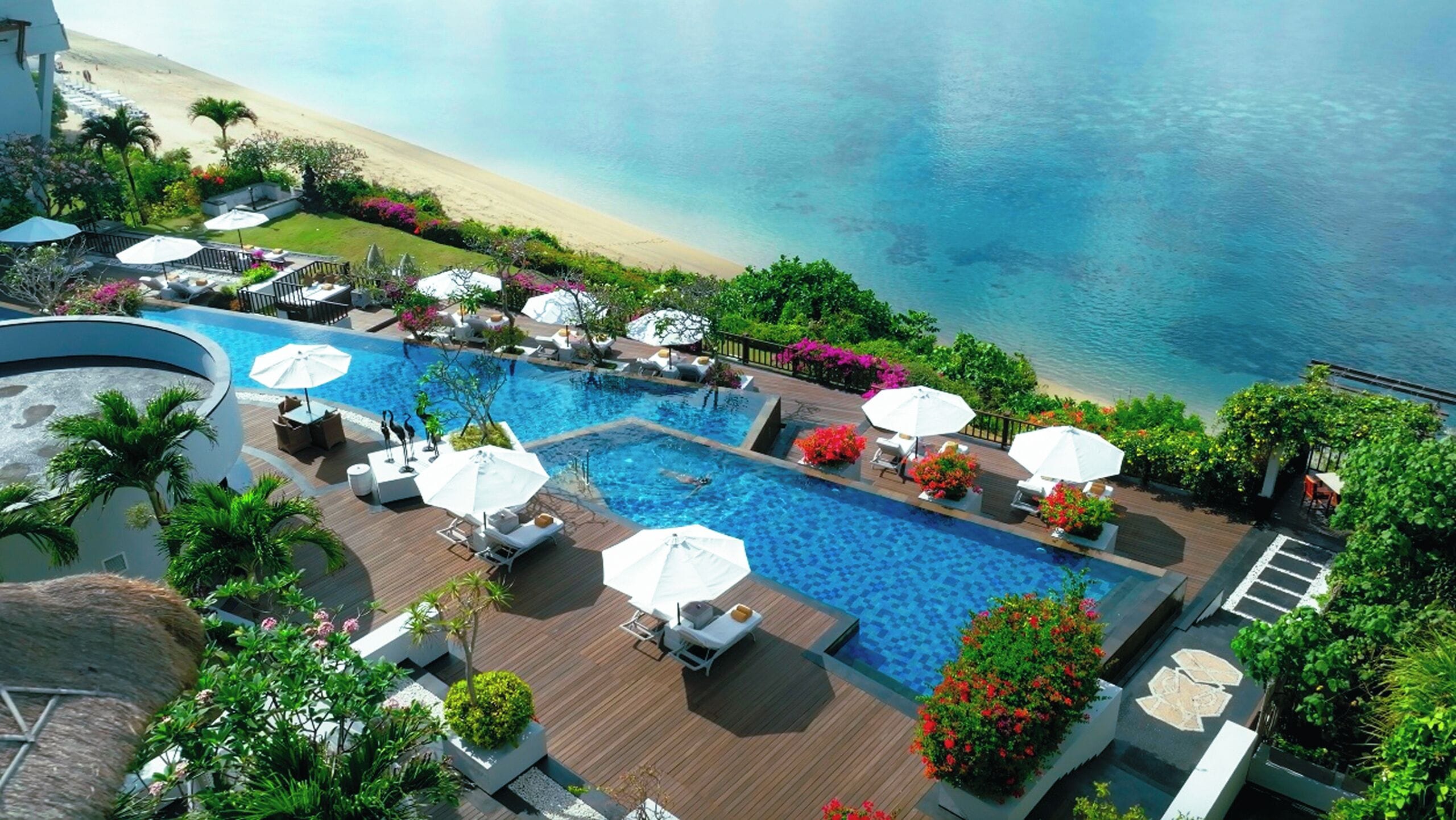 A resort pool area with multiple sun loungers and umbrellas, surrounded by lush greenery and a view of the beach.