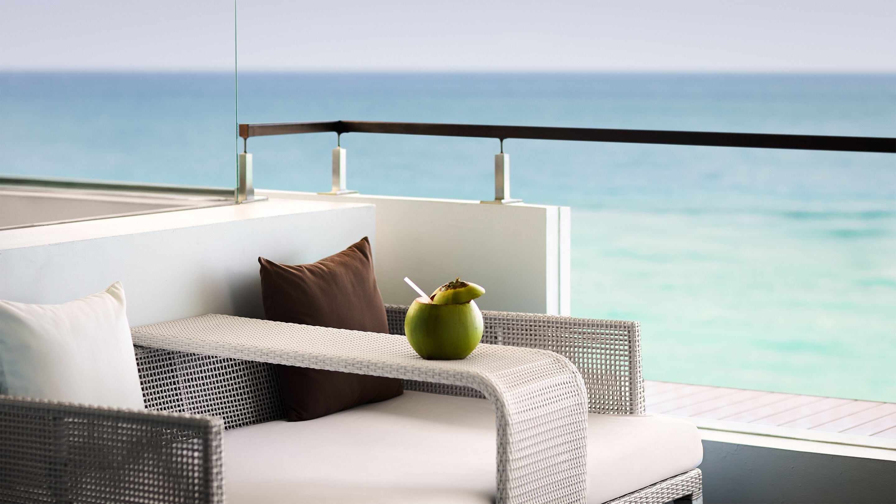 A balcony with a wicker sofa, a green vase with a straw, and a view of the ocean.