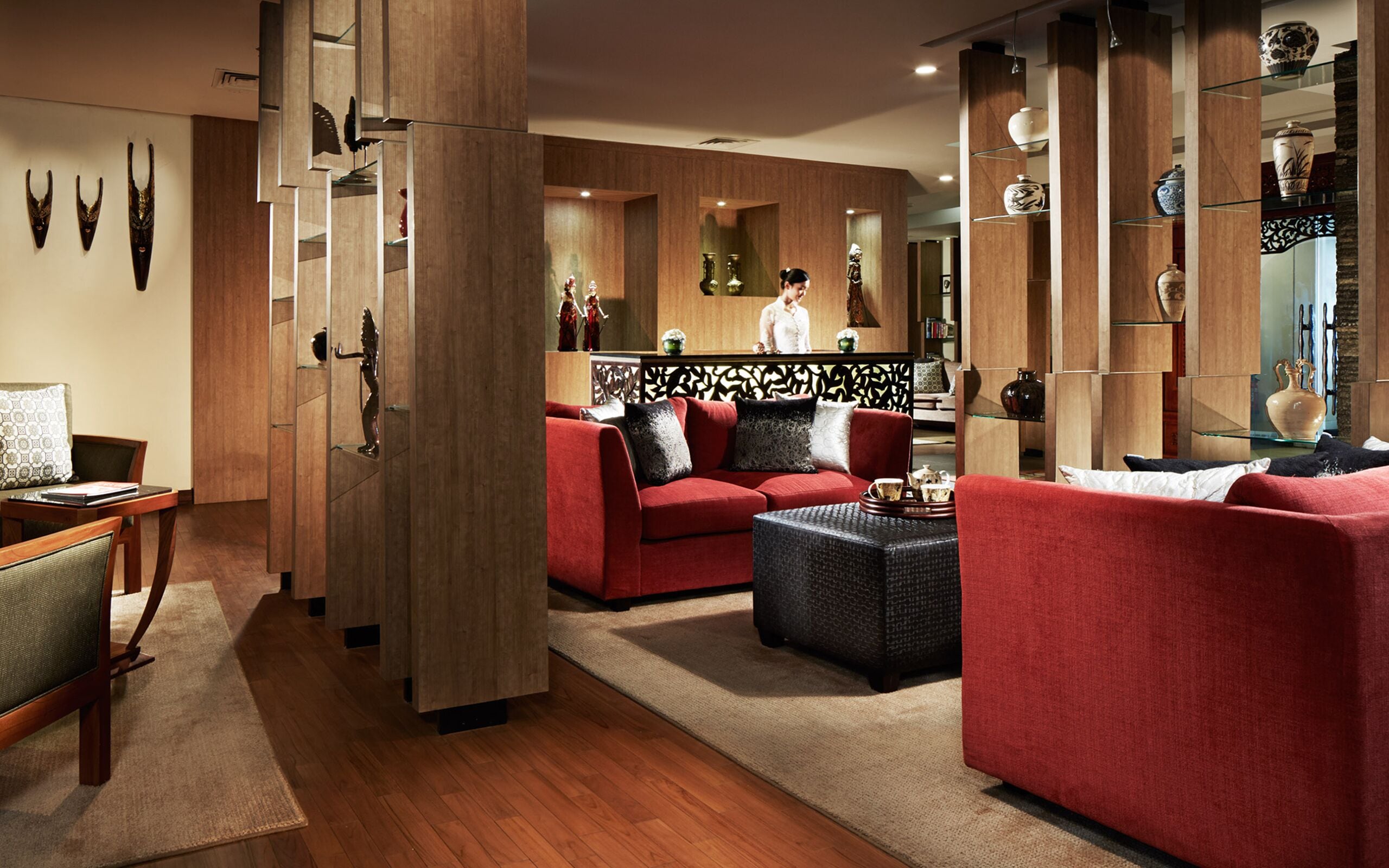 A modern living room with a red sofa, a black ottoman, and a wooden coffee table. There are decorative vases on shelves and a wooden partition with a decorative pattern.