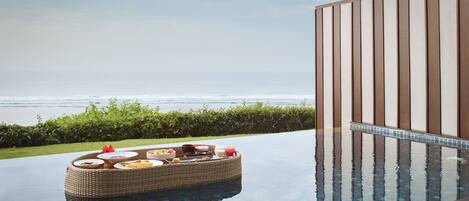 An infinity pool with a wicker lounge chair and a tray of food, overlooking the ocean.