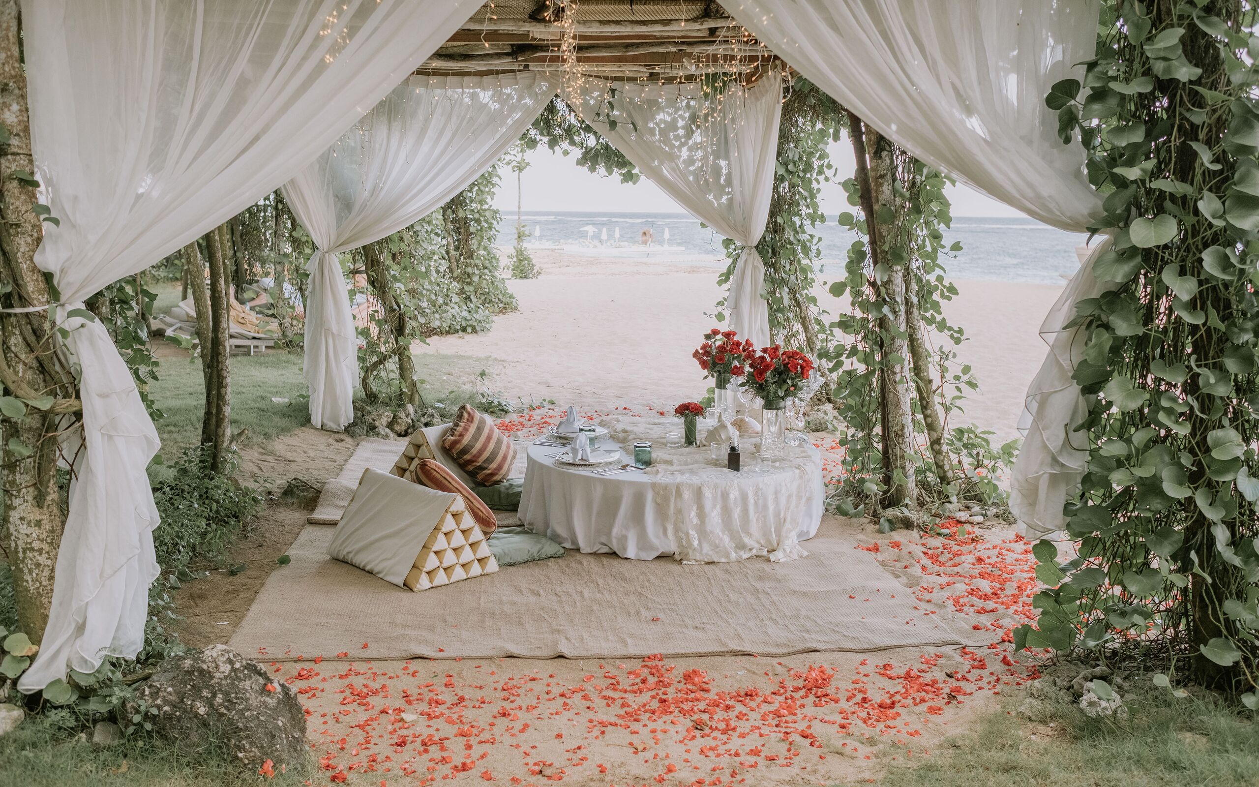 An outdoor wedding setup with a white canopy, a round table with floral arrangements, and a beach view.