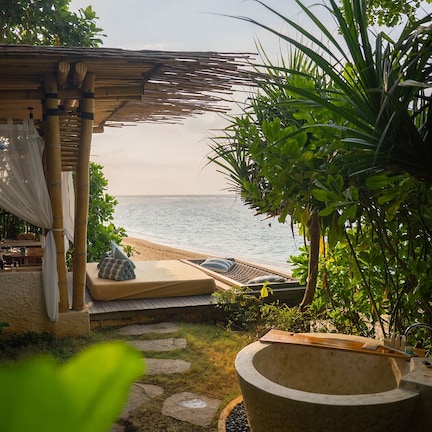 A beachside lounge area with a thatched roof, a stone fire pit, and a view of the ocean.