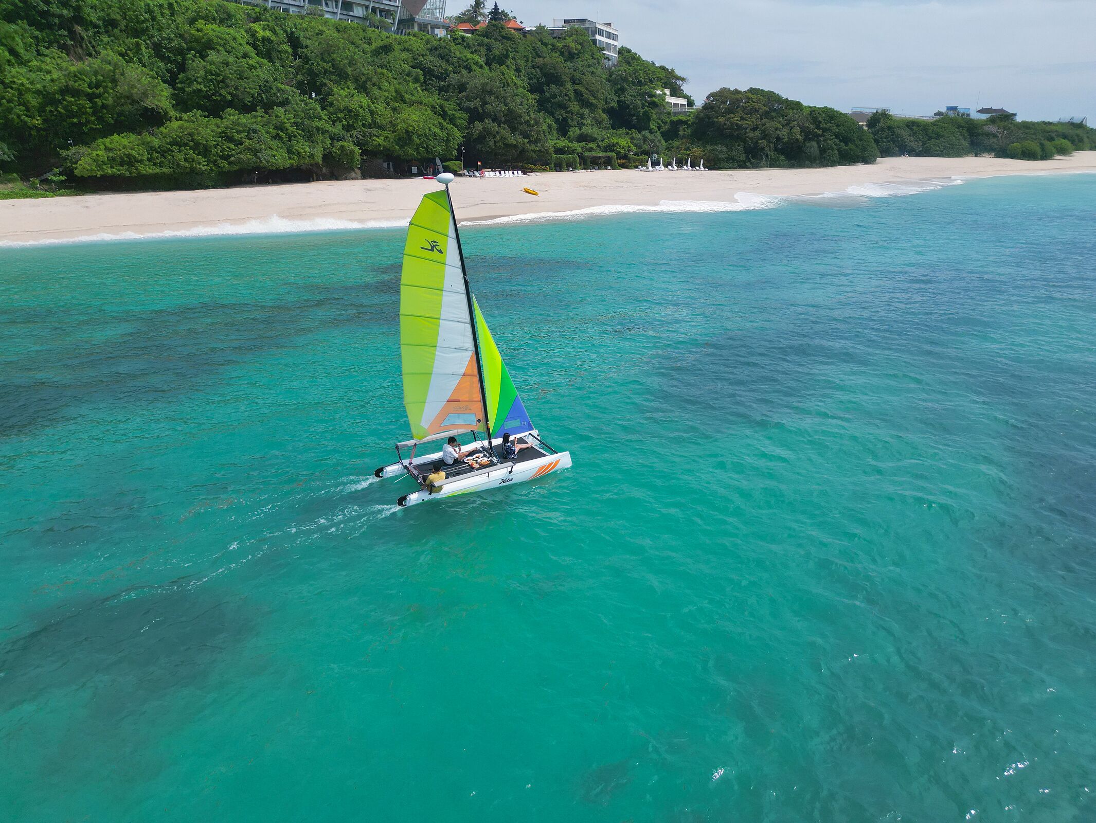 A sailboat on turquoise waters with a beach and greenery in the background.