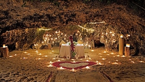 Un entorno al aire libre íntimo con un arreglo de velas en forma de corazón y una mesa con un centro de flores, todo rodeado por un ambiente tipo cueva con luces colgantes.