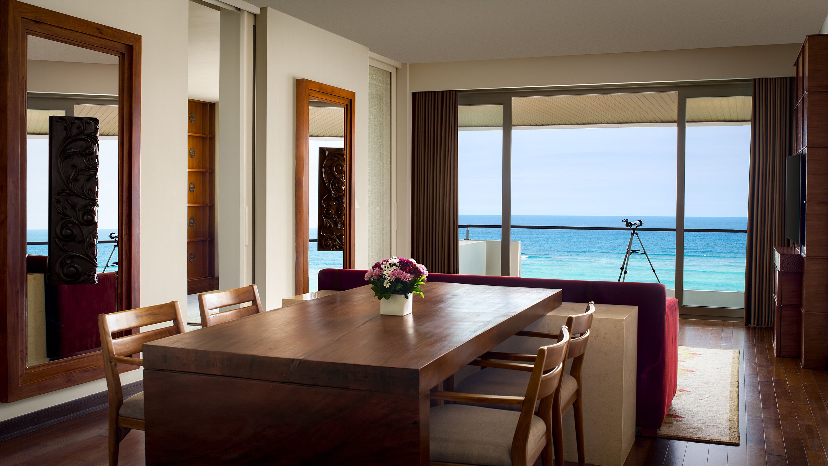 Una habitación con una mesa de comedor de madera, sillas, un pequeño arreglo floral y vista al océano a través de ventanas amplias.