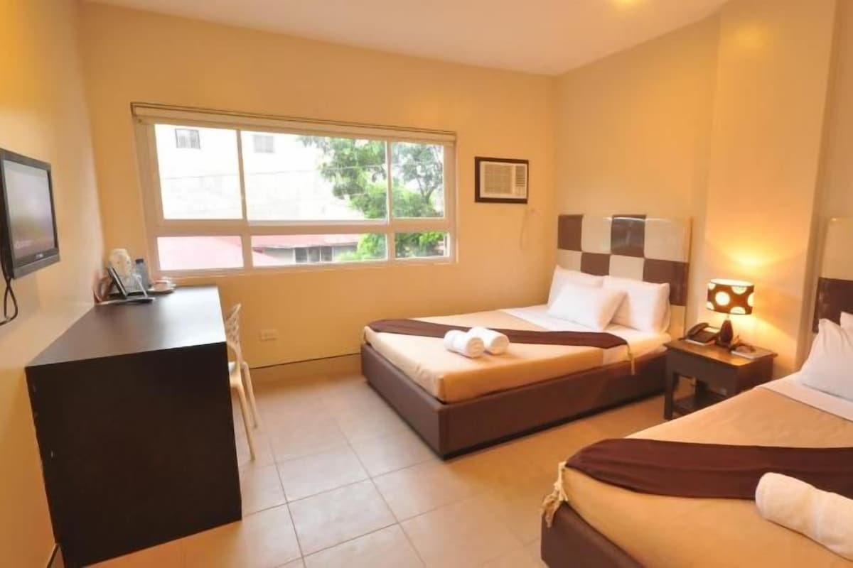Habitación de hotel con dos camas, un escritorio, un televisor y una ventana con vistas a zonas verdes.