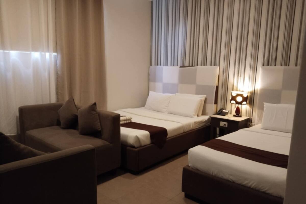 A hotel room with two beds, a sofa, a nightstand with a lamp, and curtains.