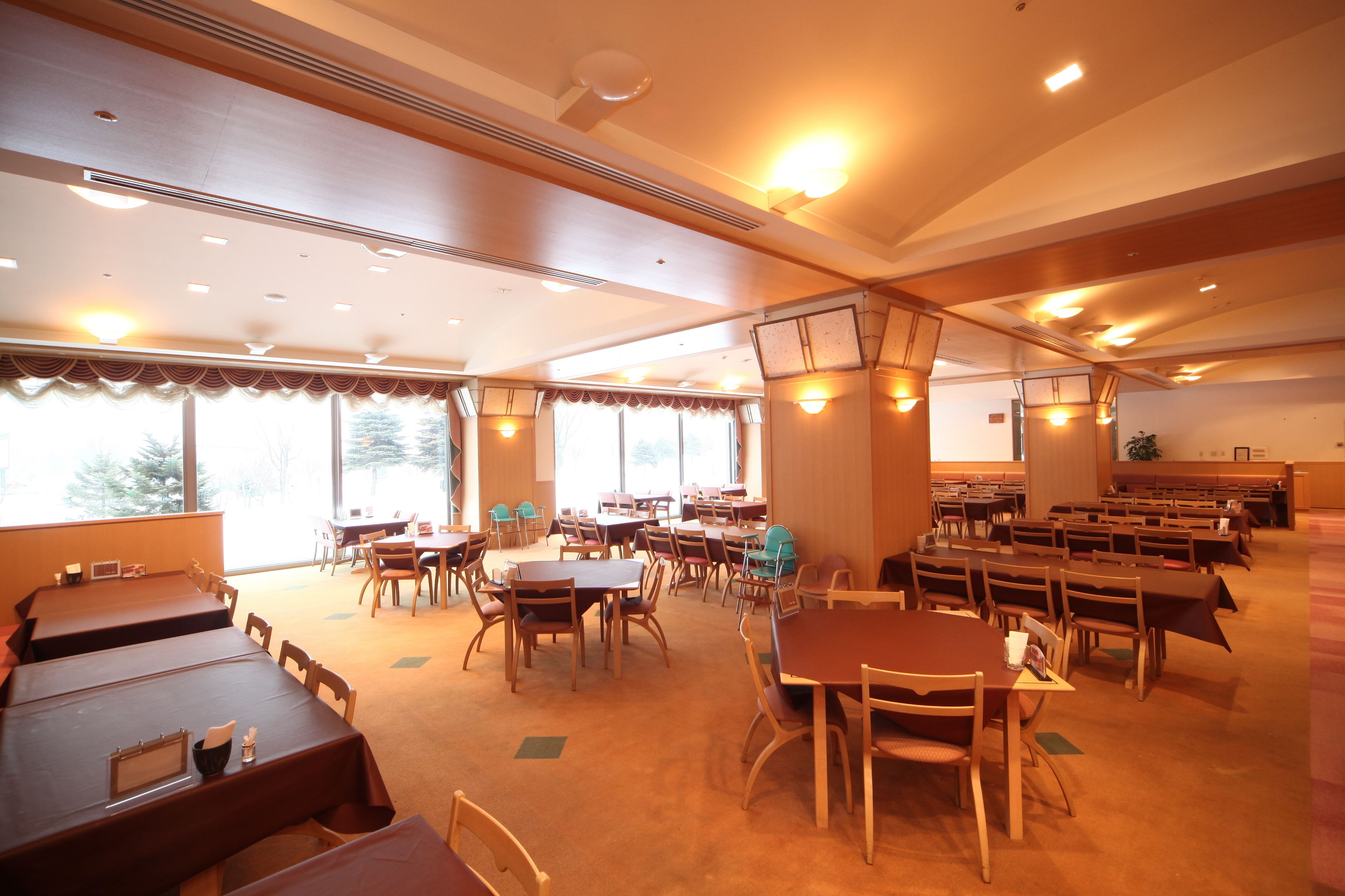 A large dining hall with multiple tables and chairs, high ceilings with recessed lighting, and large windows allowing natural light.
