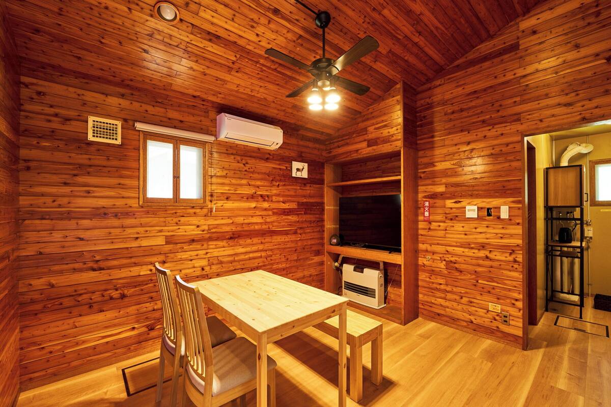 A wooden cabin interior with a dining table, chairs, a ceiling fan, an air conditioning unit, a television, and a kitchen area.
