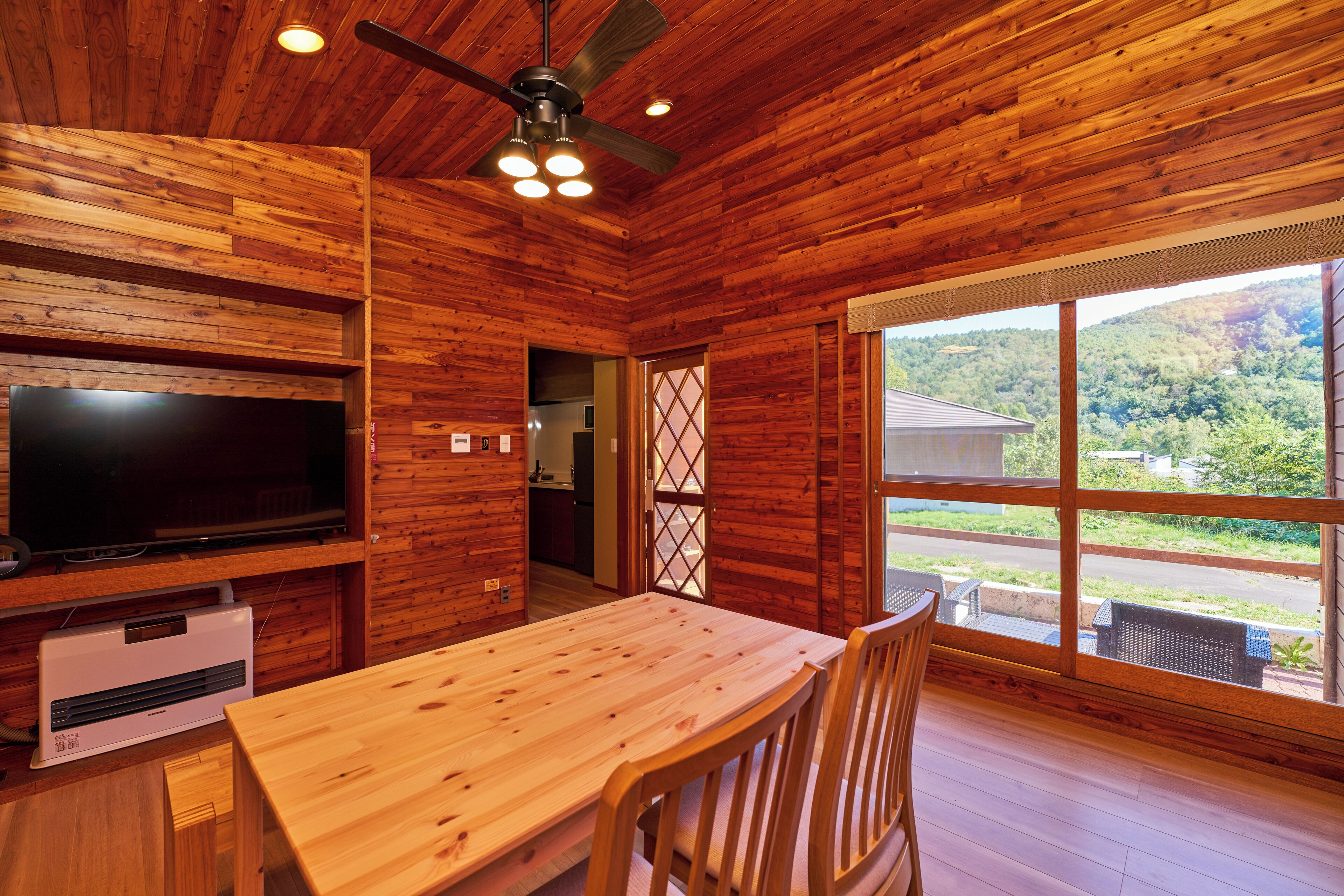 A wooden cabin interior with a dining table, chairs, a television, and a large window offering a view of the outdoors.