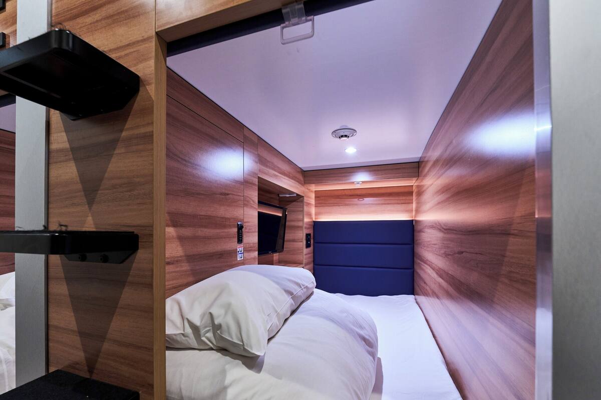 A compact hotel room with a bed, a small table, a built-in TV, and a ceiling-mounted light fixture.