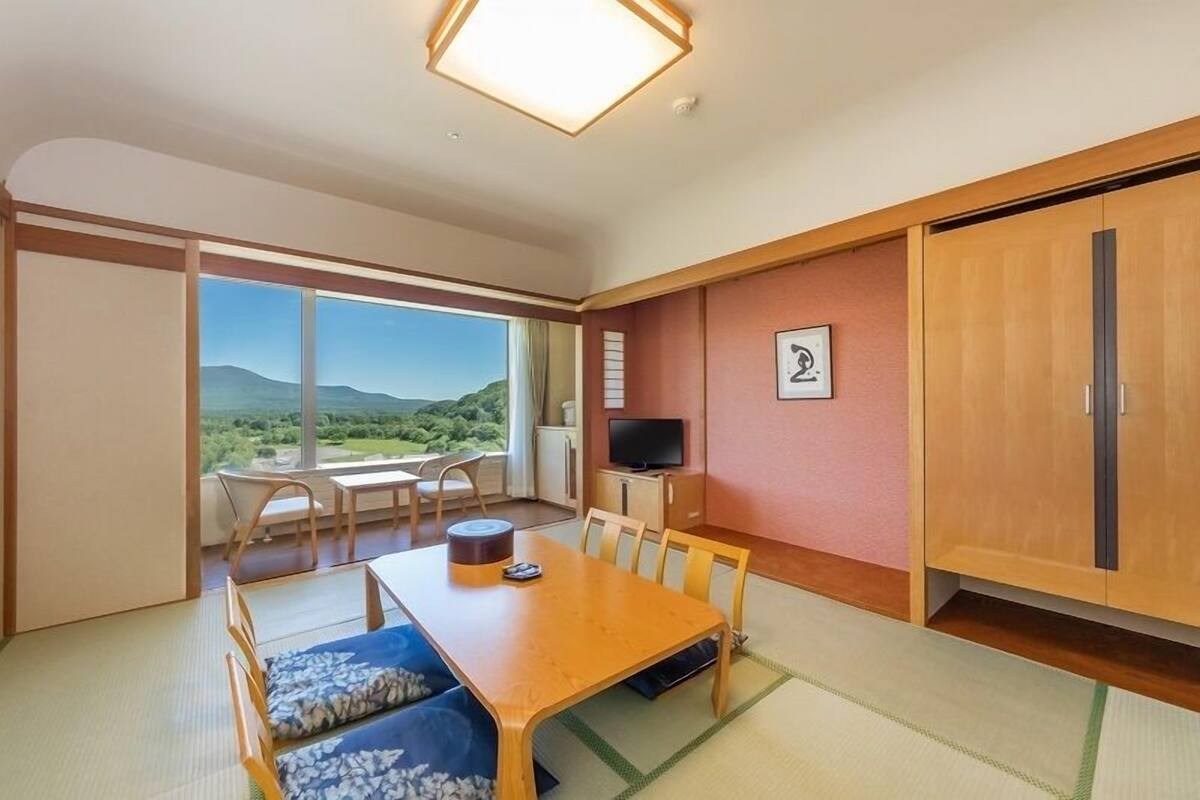 A room with tatami flooring, a wooden dining table, chairs, a television, and a sliding door leading to a balcony with a view.