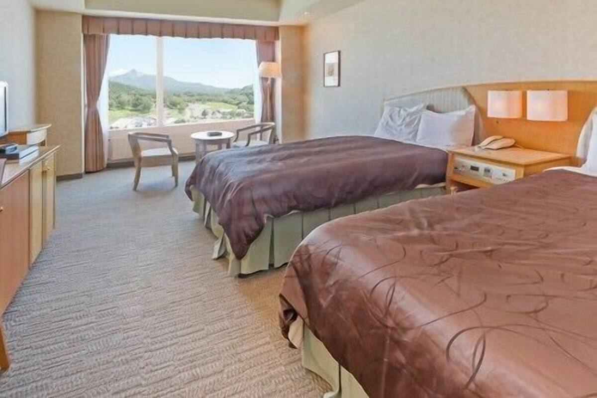 A hotel room with two beds, a desk, a chair, and a view of the outdoors.