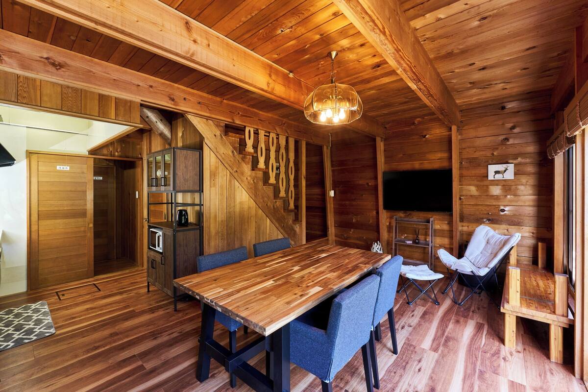A wooden cabin interior with a dining table, blue chairs, a sofa, a TV, and a staircase.