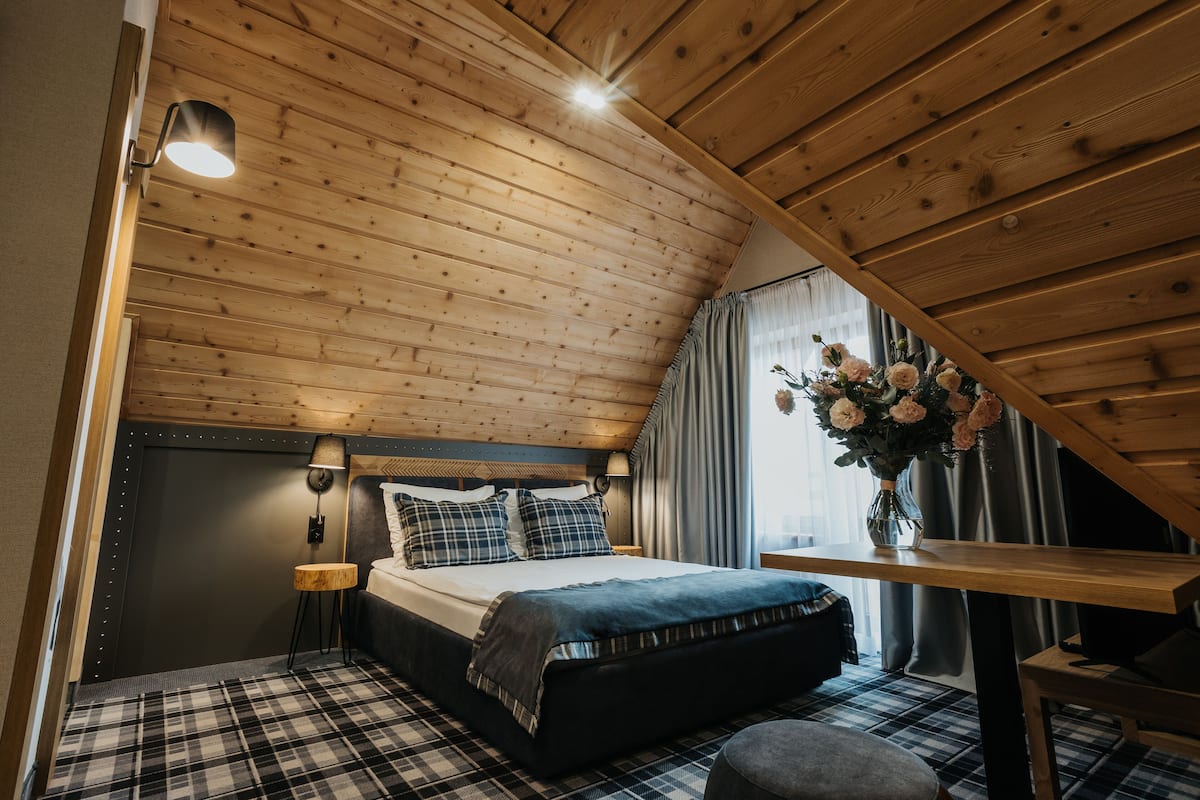 A bedroom with a wooden ceiling, a bed with a plaid bedspread, a nightstand, a lamp, and a window with curtains.