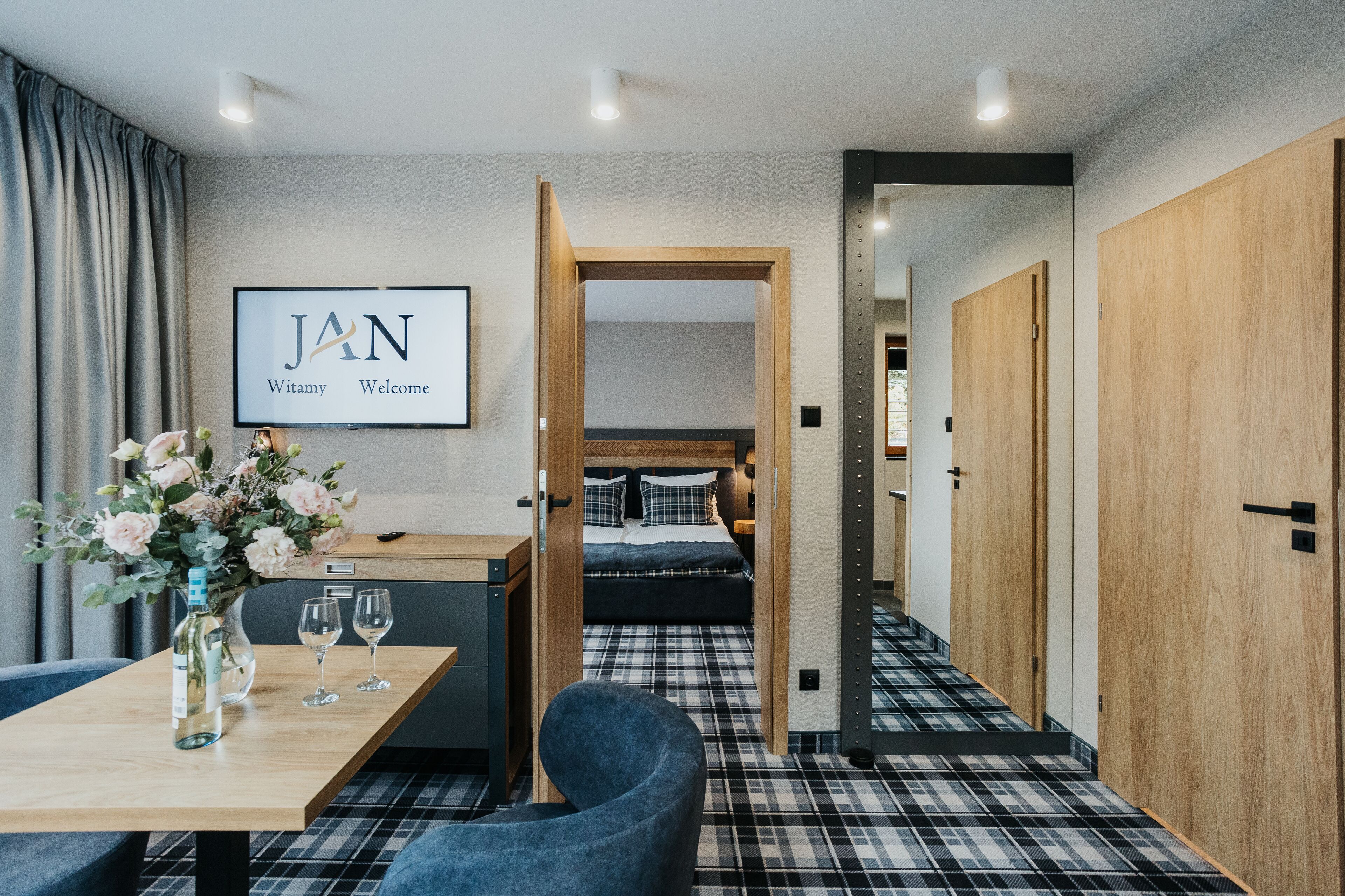 A modern hotel room with a bed, a desk, a chair, and a TV displaying "JAN Welcome".