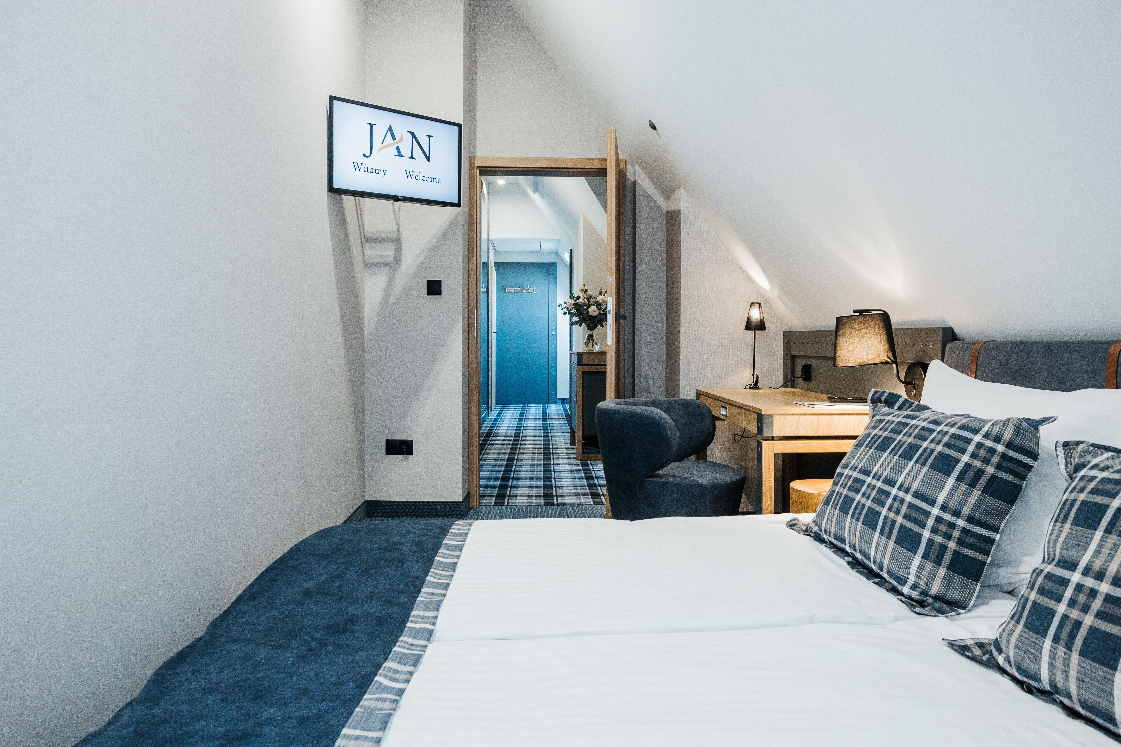 Quarto de hotel com cama, escrivaninha, cadeira, abajur e placa com a mensagem "JAN Bem-vindo".