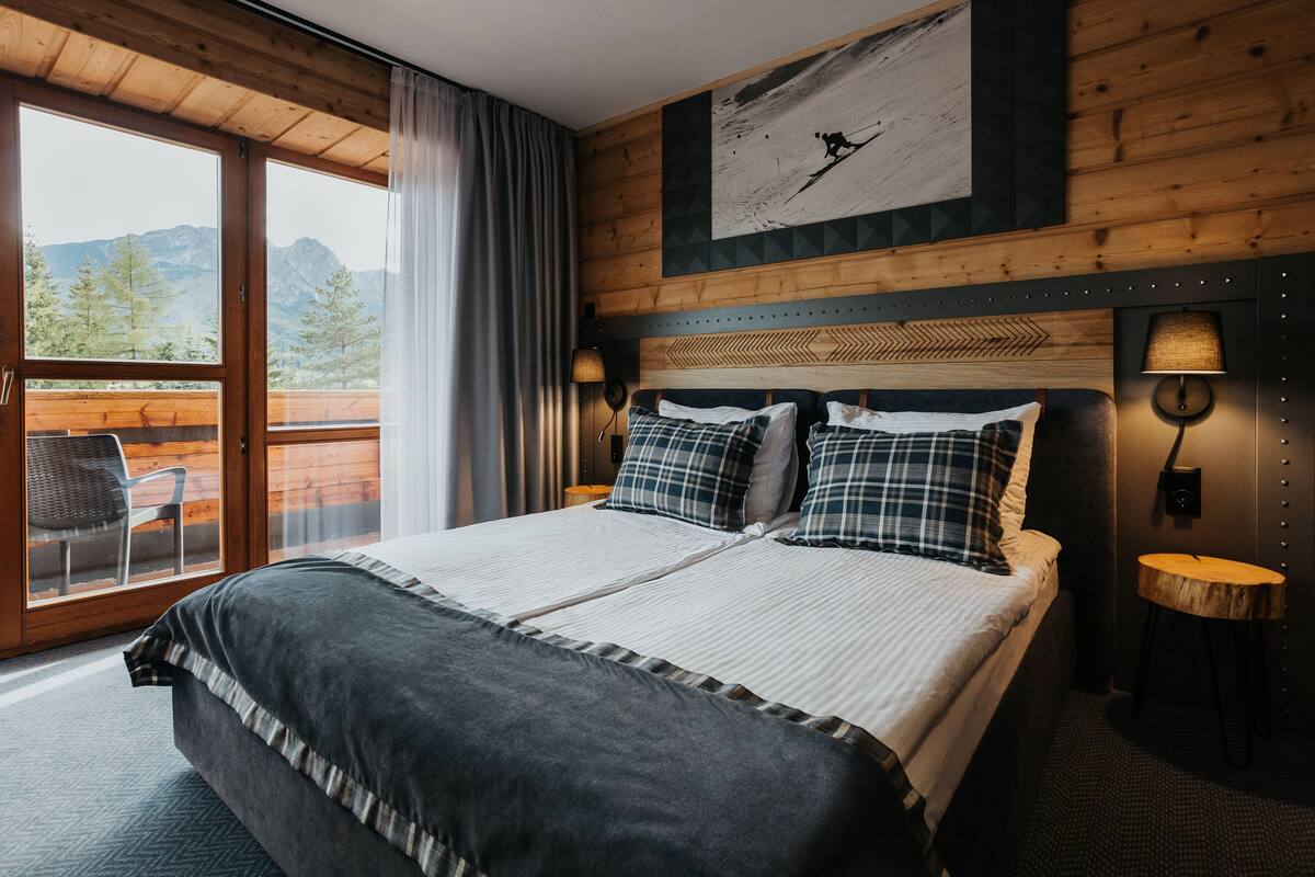 A bedroom with a large bed, two bedside lamps, a wooden headboard, and a view of mountains through a sliding door.
