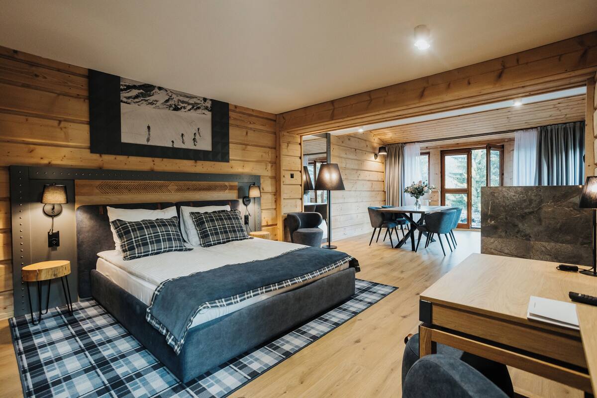 A modern bedroom with a large bed, bedside lamps, a wooden headboard, a dining area with a table and chairs, and a view of the outdoors through a window.