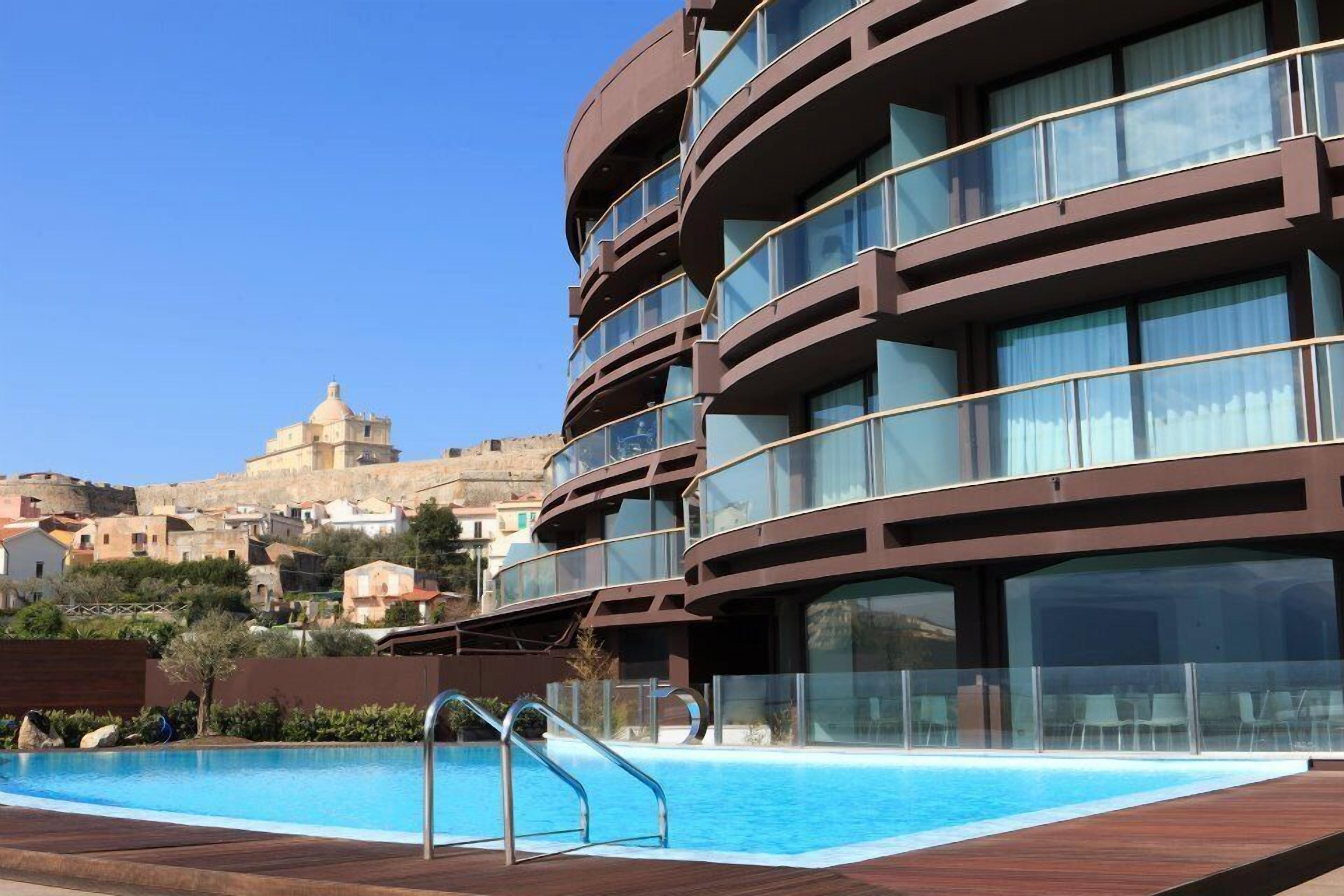 Ett modernt hotell med pool, stora fönster och utsikt över en historisk byggnad i bakgrunden.
