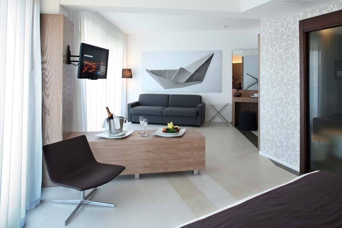 A modern hotel room with a sofa, a wooden coffee table, a TV mounted on the wall, and a large abstract painting.