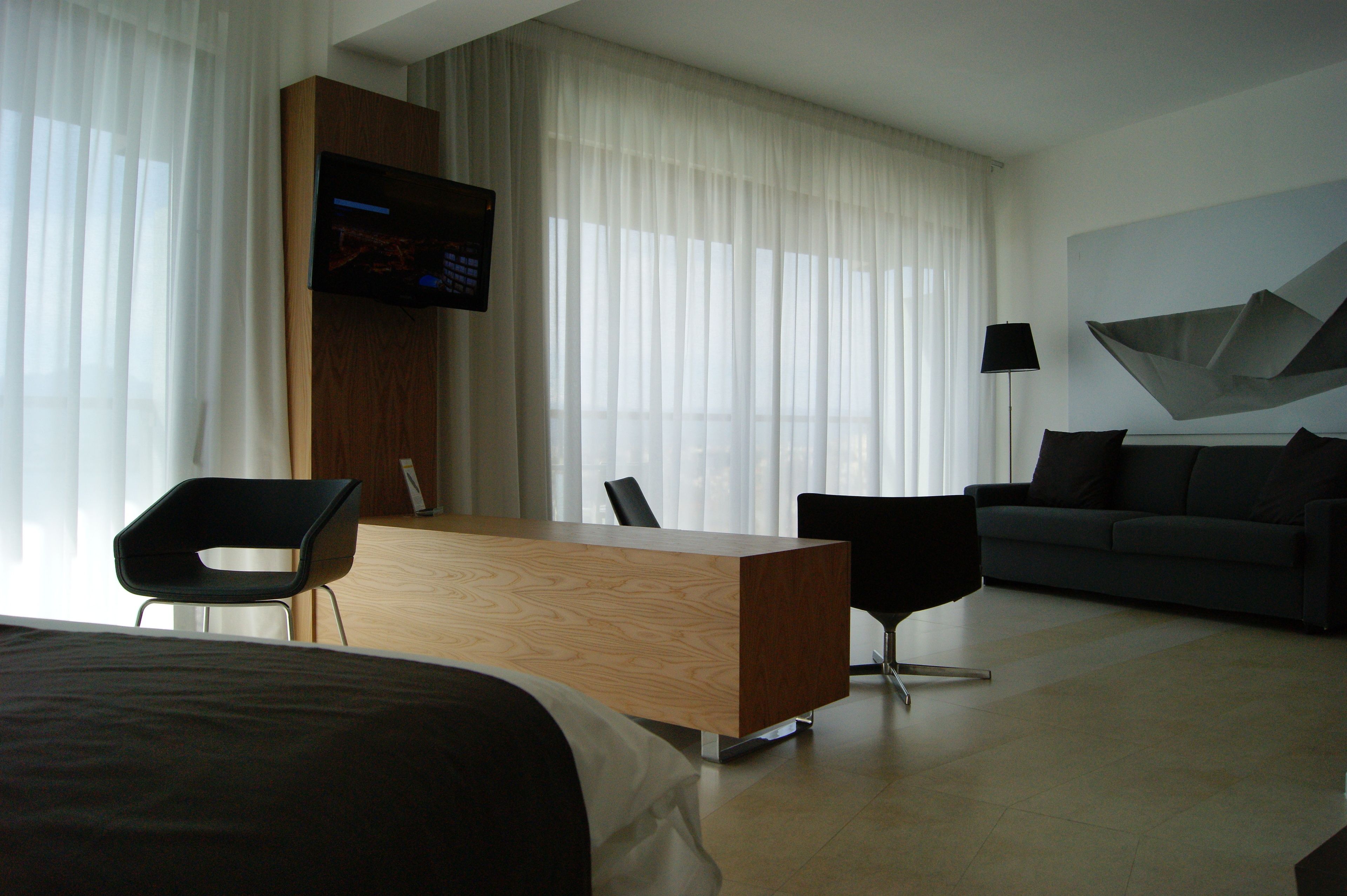 A modern hotel room with a wooden desk, two black chairs, a grey sofa, a flat-screen TV mounted on the wall, and sheer curtains.