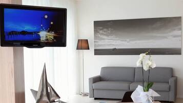 Una sala de estar moderna con un televisor de pantalla plana, un sofá gris, una mesa de centro con un libro y un jarrón de orquídeas blancas, y un cuadro de paisaje grande en la pared.