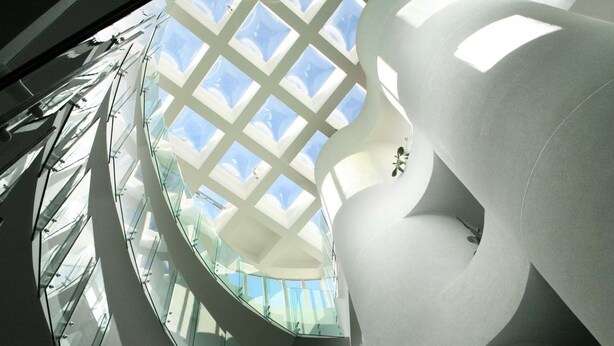 Un intérieur architectural moderne avec un plafond en verre, des murs blancs aux lignes courbes et un escalier en colimaçon.