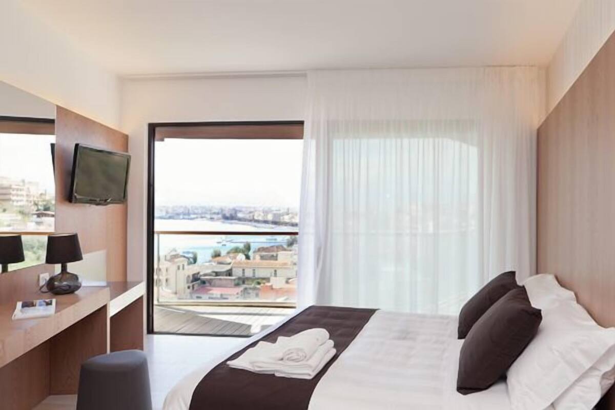 A hotel room with a bed, a desk with a laptop, a television, and a view of the city through large windows.