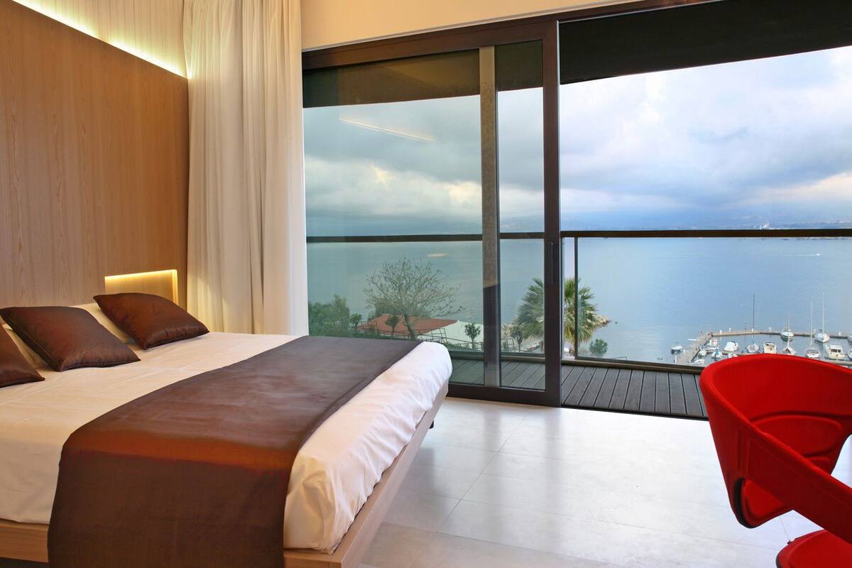 A bedroom with a large bed, a red chair, and a view of the sea and marina.