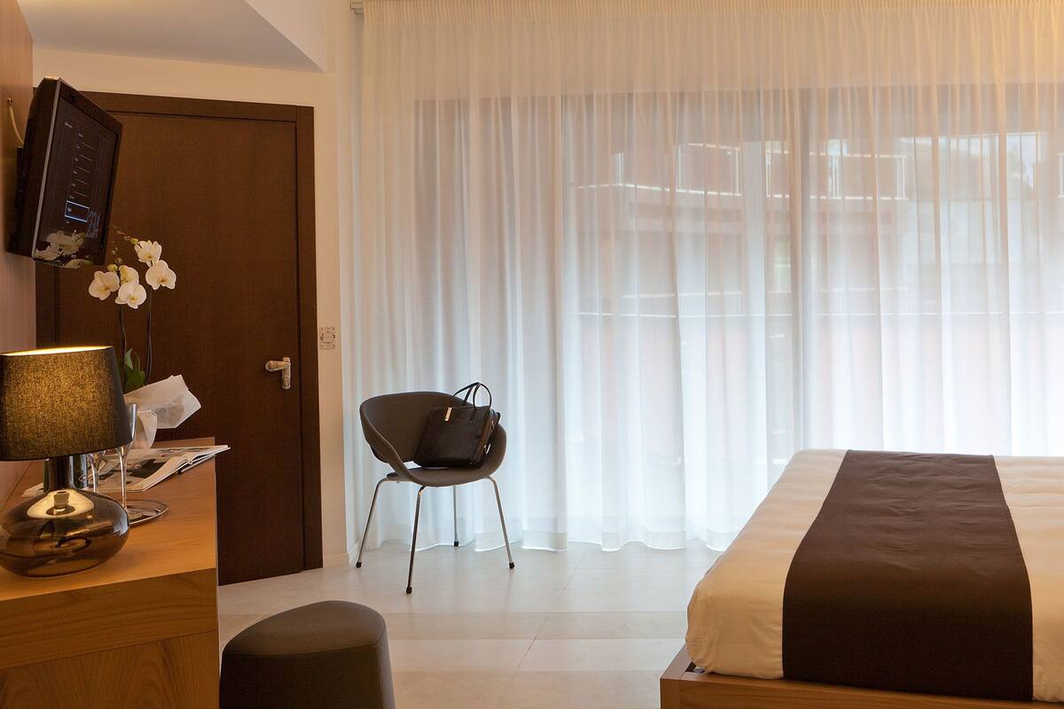 A hotel room with a bed, a chair, a desk with a lamp, and a TV on the wall.