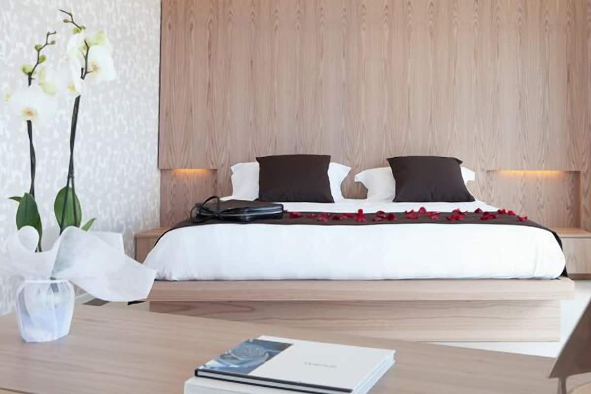 A modern bedroom with a wooden headboard, a neatly made bed with white and black bedding, and a bedside table with a book and a vase of flowers.