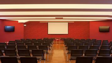 Uma sala de conferências com fileiras de cadeiras, uma tela de projetor e uma televisão na parede.