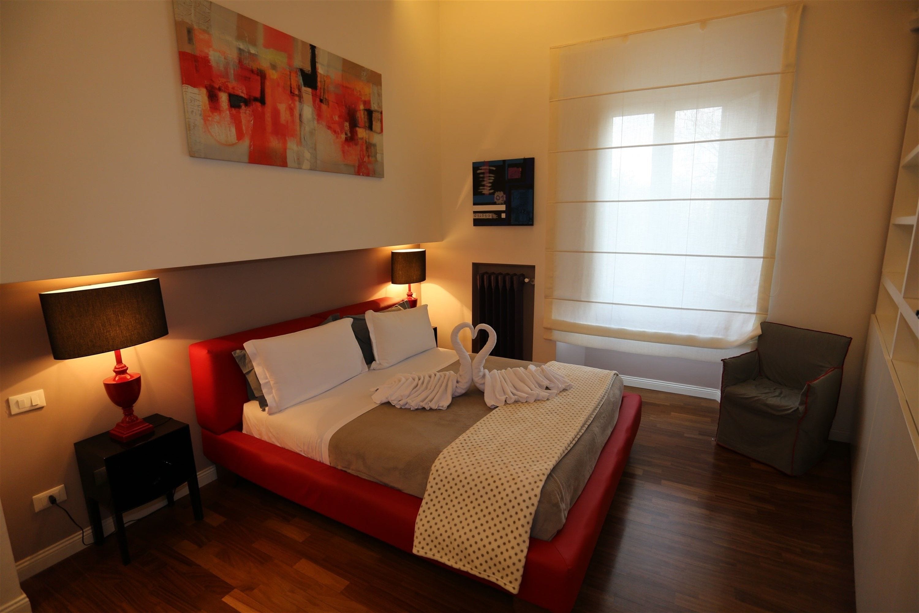 A modern hotel room with a red bed, two bedside lamps, a large window with blinds, and a red chair.