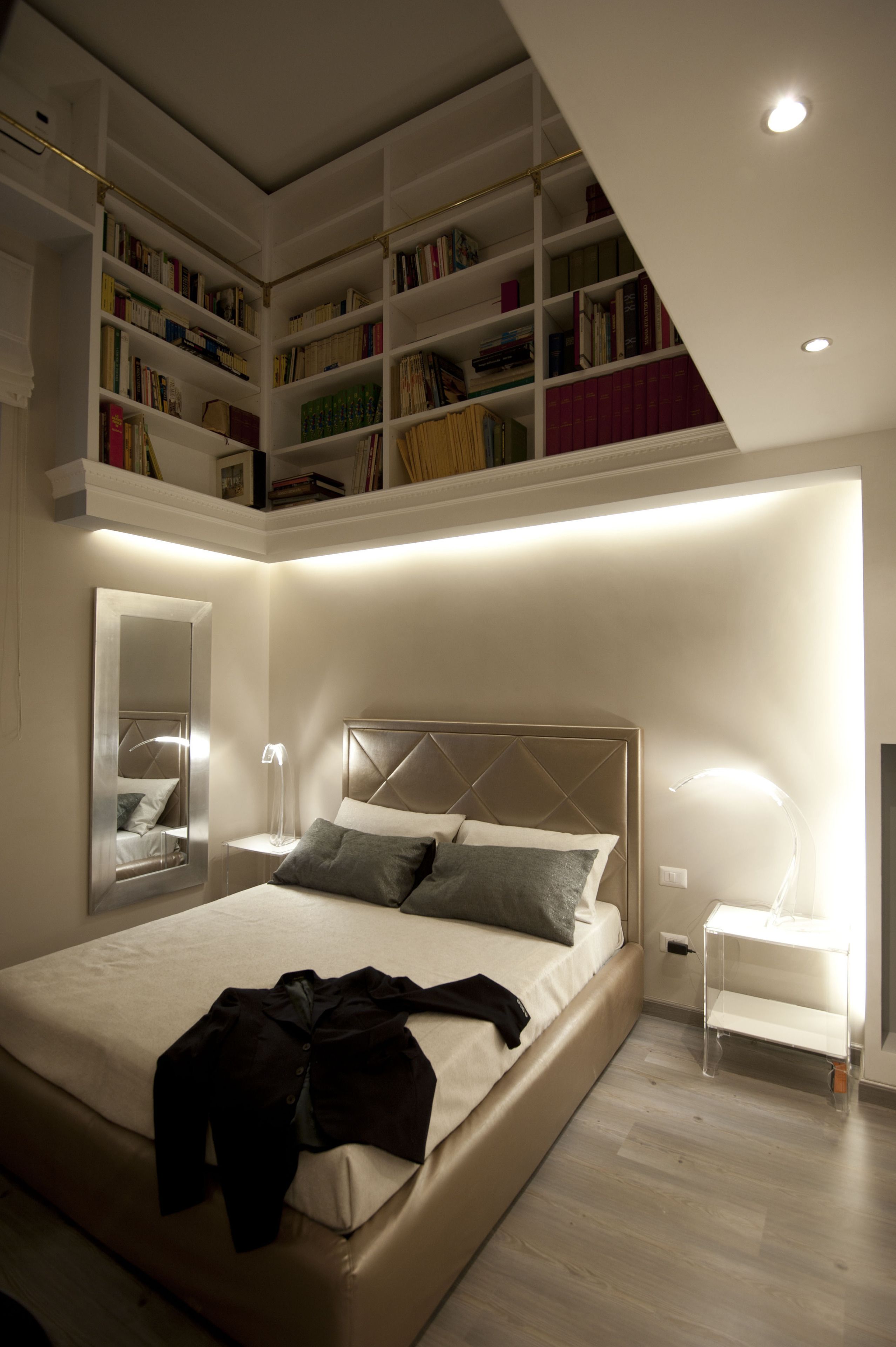 A modern bedroom with a large bed, built-in bookshelves, a mirror, and a bedside table.
