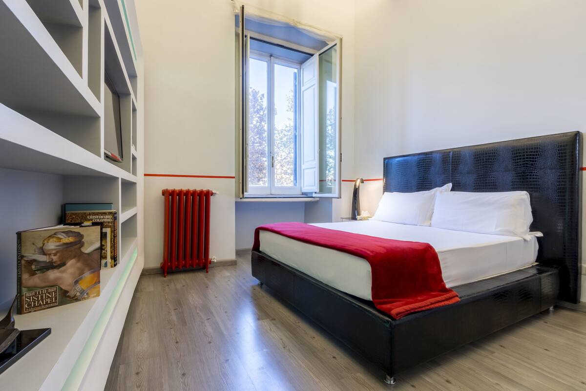 A modern bedroom with a black headboard, white bedding, a red throw blanket, and a bookshelf with books.