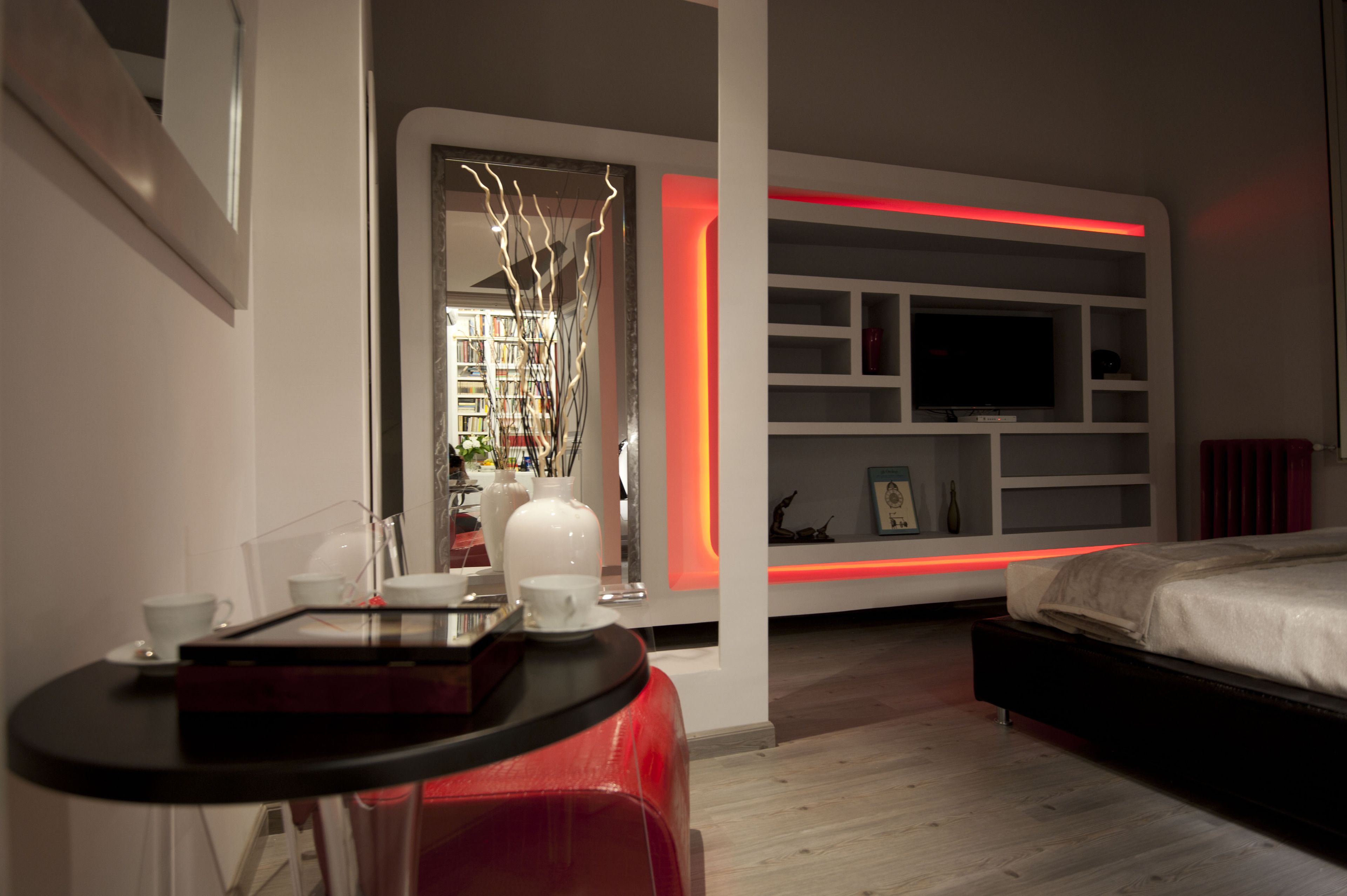 A modern hotel room with a bed, a TV, a bookshelf, and a small table with a cup and saucer.