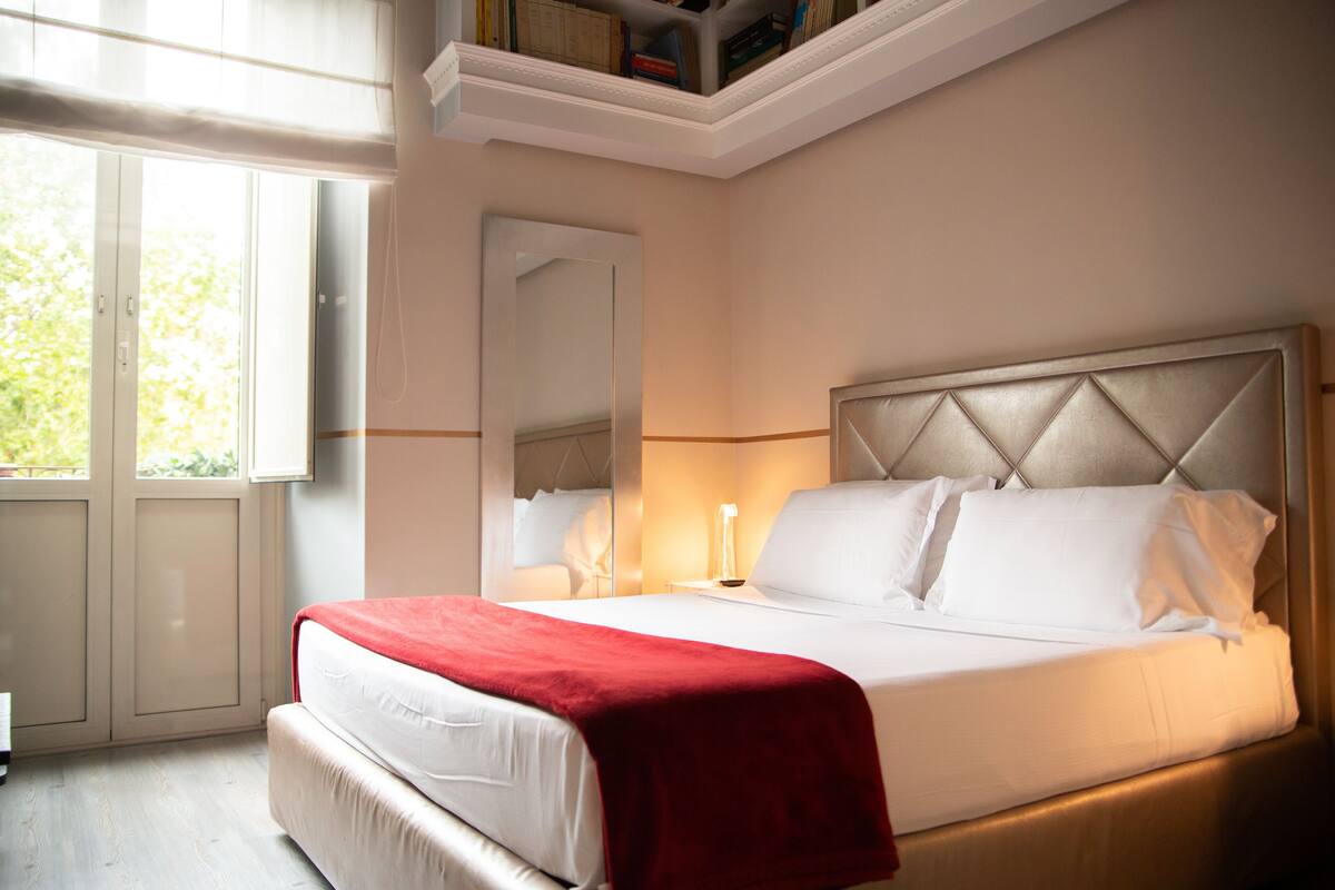 A neatly made bed with a red blanket, a bedside lamp, and a large mirror on the wall.