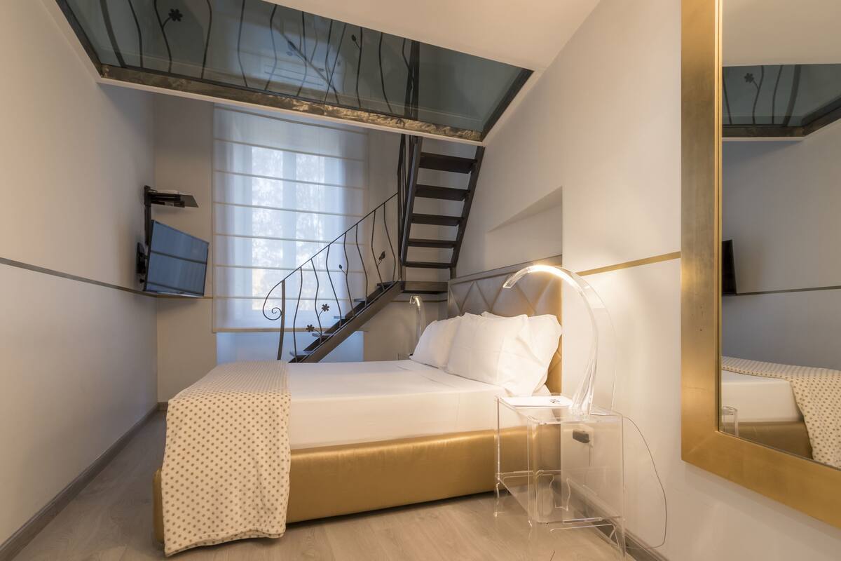 A bedroom with a bed, a bedside table, a lamp, a mirror, and a staircase leading to an upper level.