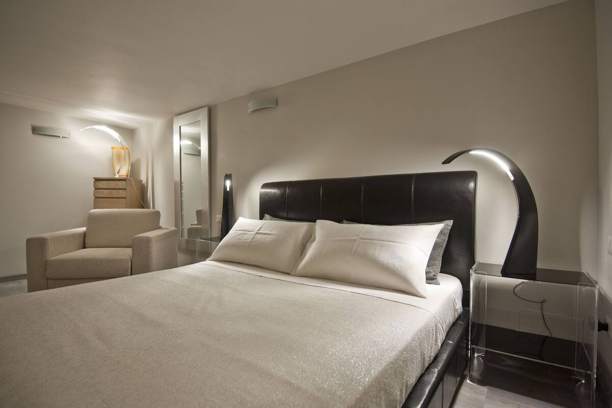 A modern bedroom with a large bed, a chair, a bedside table, and a lamp.