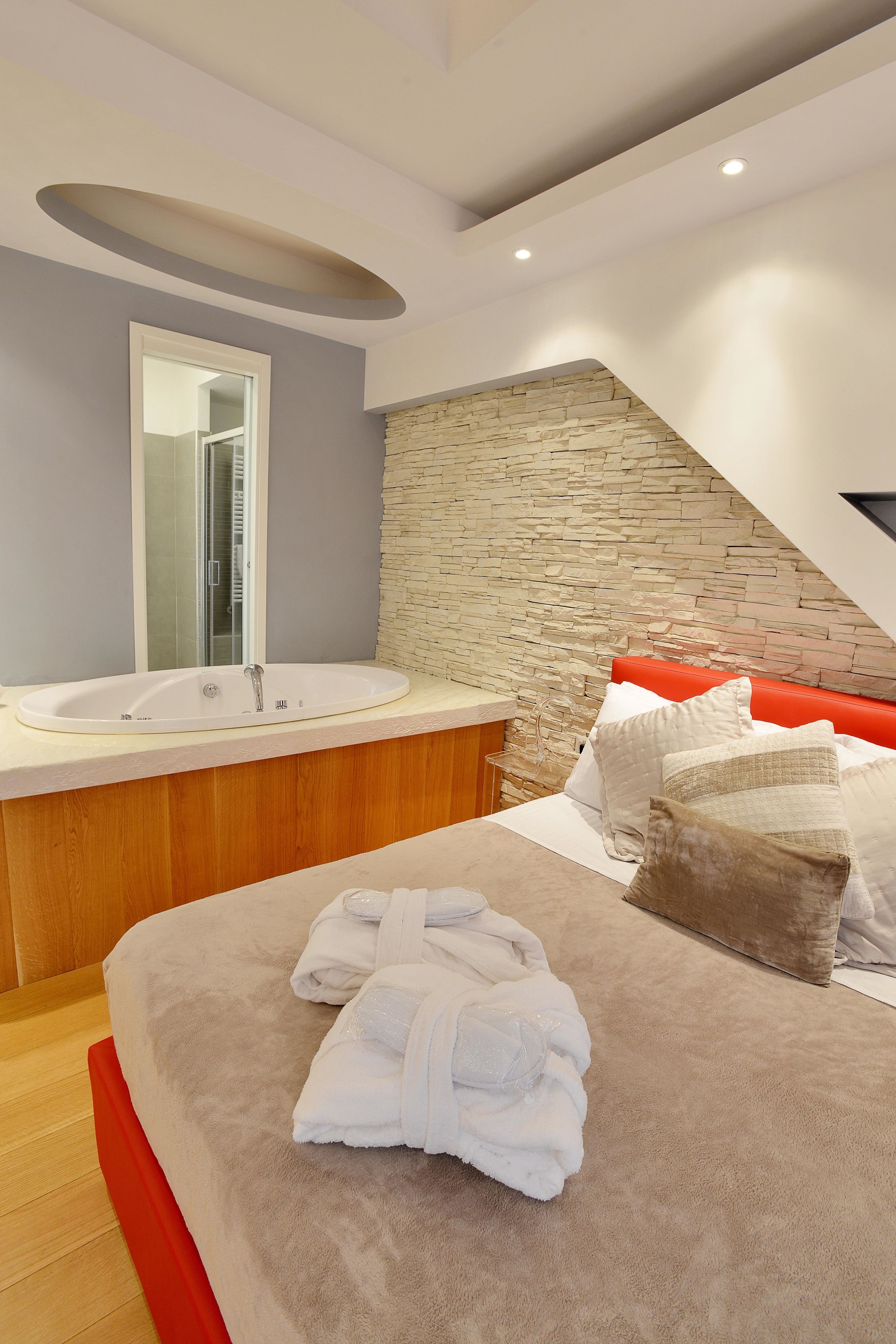 Een moderne slaapkamer met een accentmuur van steen, een groot bed met wit en bruin beddengoed, een houten hoofdbord, en een badkamer met een betegelde muur en een grote spiegel.