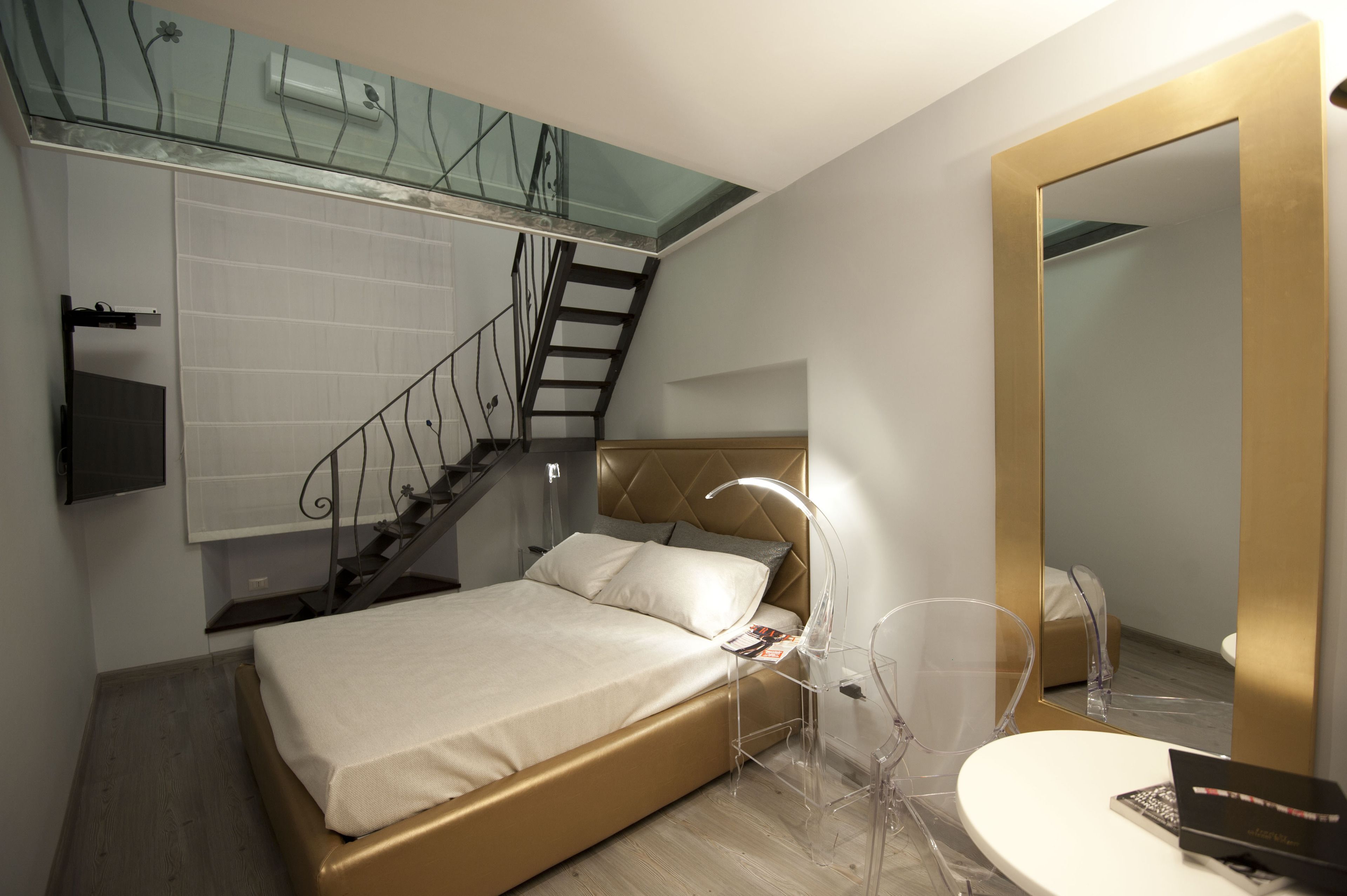 Un dormitori modern amb un llit, una escala de vidre, una taula de nit, una cadira i un mirall de bany.